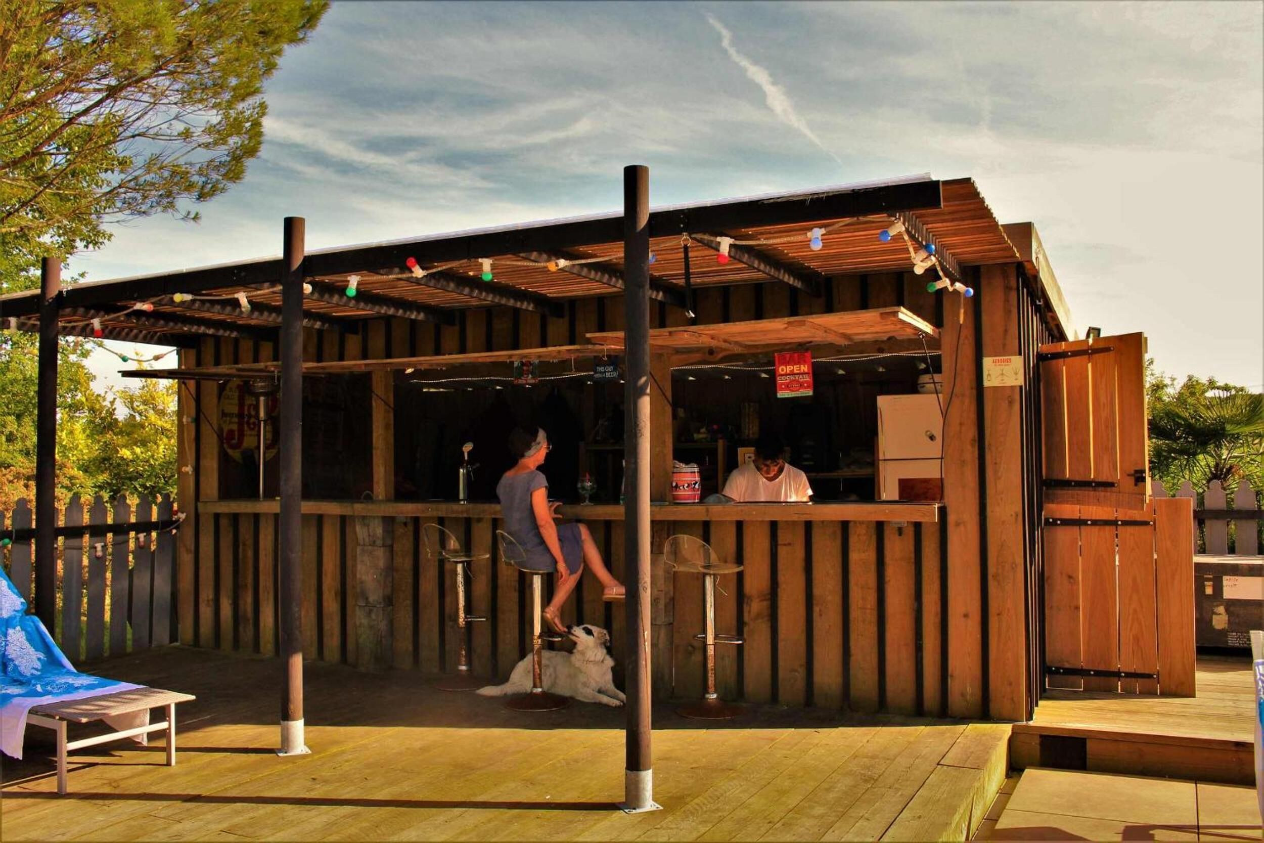 Un bar extérieur en bois doté d’un espace salon couvert, une personne debout au bar et un chien allongé sur le sol.