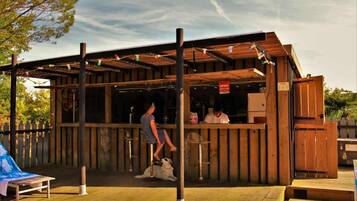 Un bar extérieur en bois doté d’un espace salon couvert, une personne debout au bar et un chien allongé sur le sol.