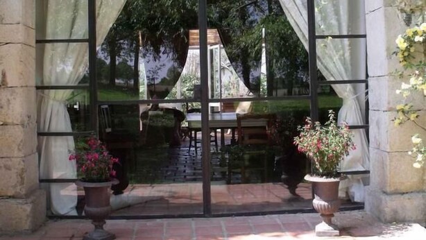 An arched entrance with a glass door and curtains, leading to an outdoor seating area with tables and chairs, surrounded by potted plants. - Maison Basta (Orthevielle)
