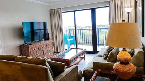 Ein Wohnzimmer mit Fernseher, Korbmöbeln, einem Glastisch und einem Balkon mit Aussicht.