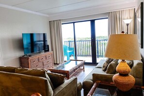 Una sala de estar con televisor, muebles de mimbre, una mesa de centro de vidrio y un balcón con vistas.