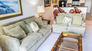 Ein Wohnzimmer mit zwei grauen Sofas, einem Korbstuhl, einer Lampe und einer Küche im Hintergrund.