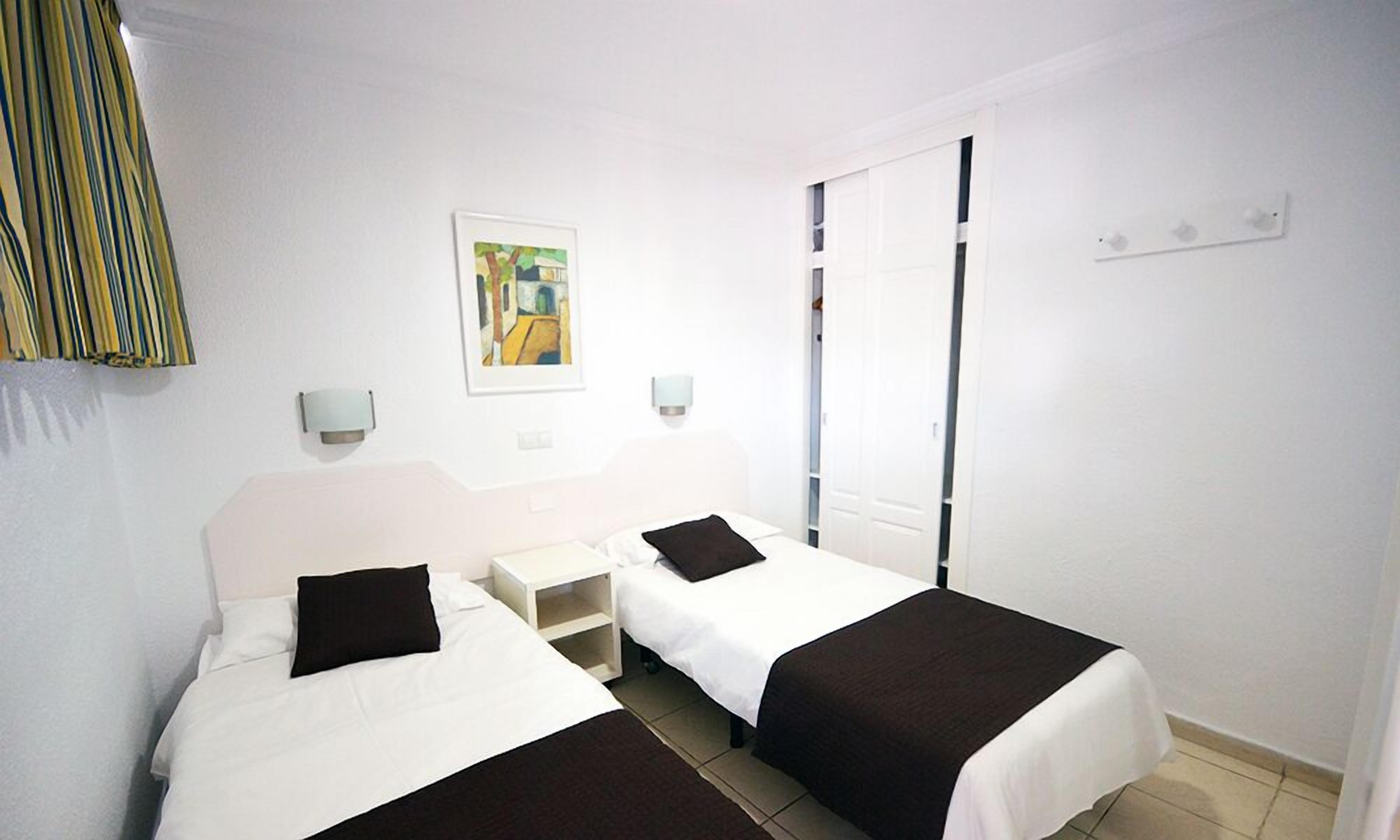 A hotel room with two beds, a nightstand, a painting, and a window with blinds.