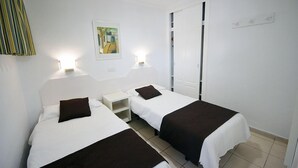 A twin room with two beds, each with a pillow and a nightstand. There is a painting on the wall and a window with a view. - Apartamentos Arco Iris (San Bartolomé de Tirajana)
