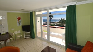 A living room with a television, a dining area with a table and chairs, and a balcony with a view of the beach. - Apartamentos Arco Iris (San Bartolomé de Tirajana)