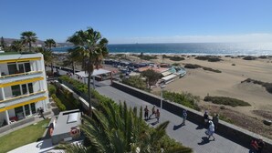 A coastal promenade with a beach, palm trees, and a modern building with yellow accents. - Apartamentos Arco Iris (San Bartolomé de Tirajana)