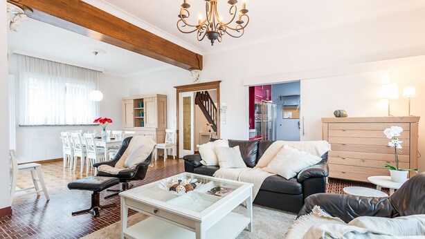 A spacious living room with a chandelier, a dining area, and a kitchen visible through an open door. - Marsouins (Koksijde)