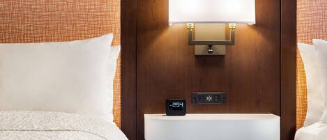 Habitación de hotel con cama, mesita de noche con reloj digital y lámpara fijada a la pared.