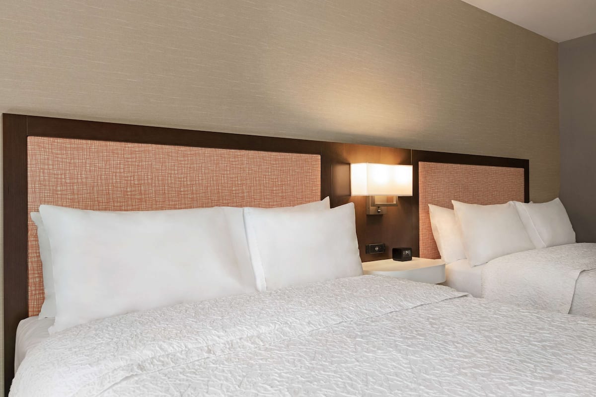 A hotel room with two beds, a nightstand, a lamp, and a wall-mounted mirror.