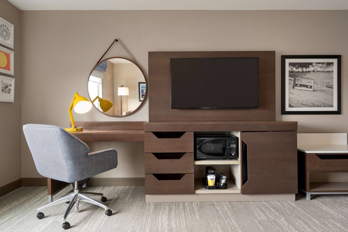 A modern hotel room with a desk, chair, flat-screen TV, microwave, and a mirror.