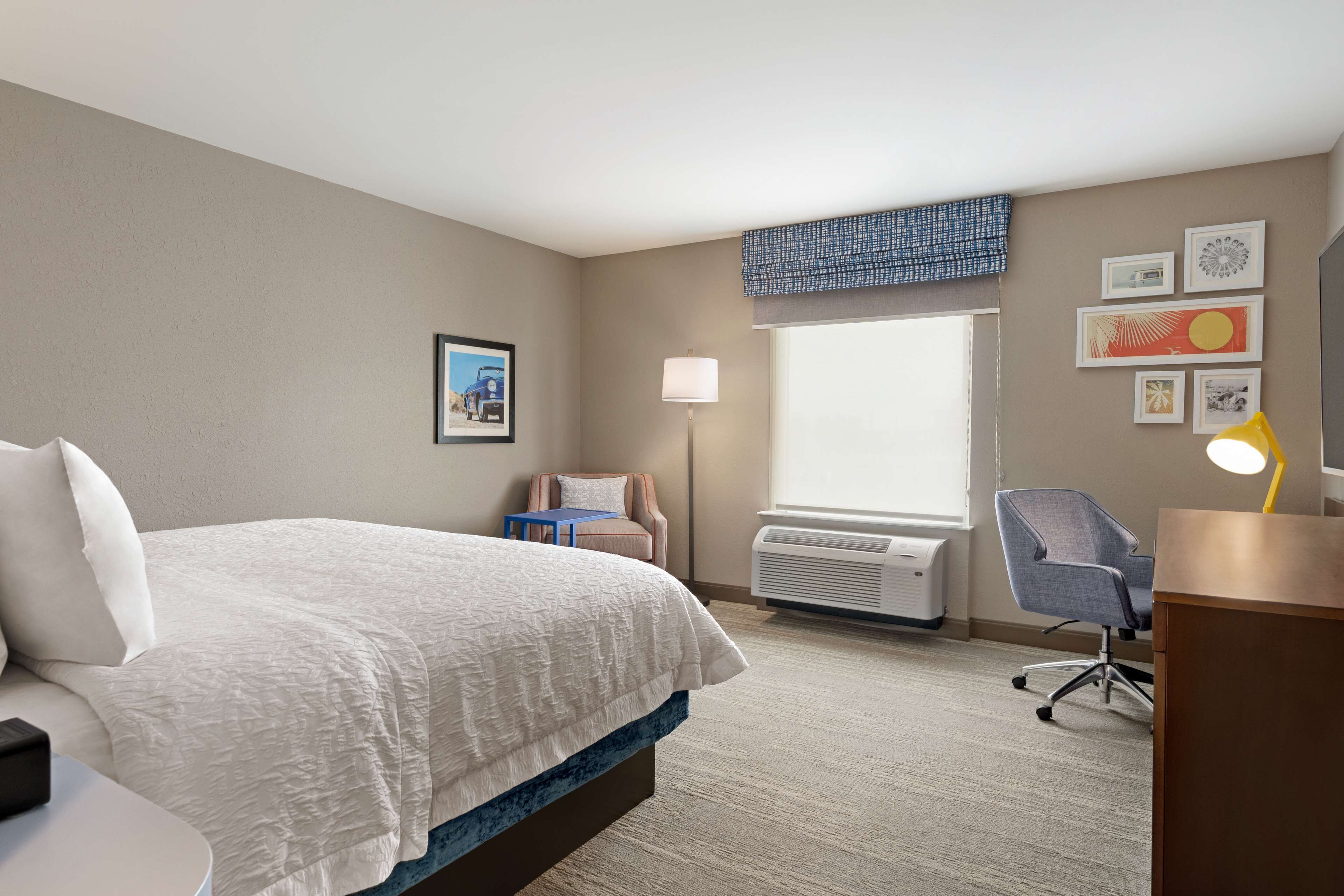 A hotel room with a bed, a desk, a chair, a lamp, and a window with blinds.