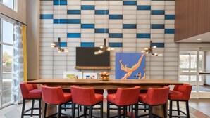 A modern dining area with a long wooden table, red upholstered chairs, and a blue and white patterned wall. - Hampton Inn & Suites Ocean City West (Ocean City)