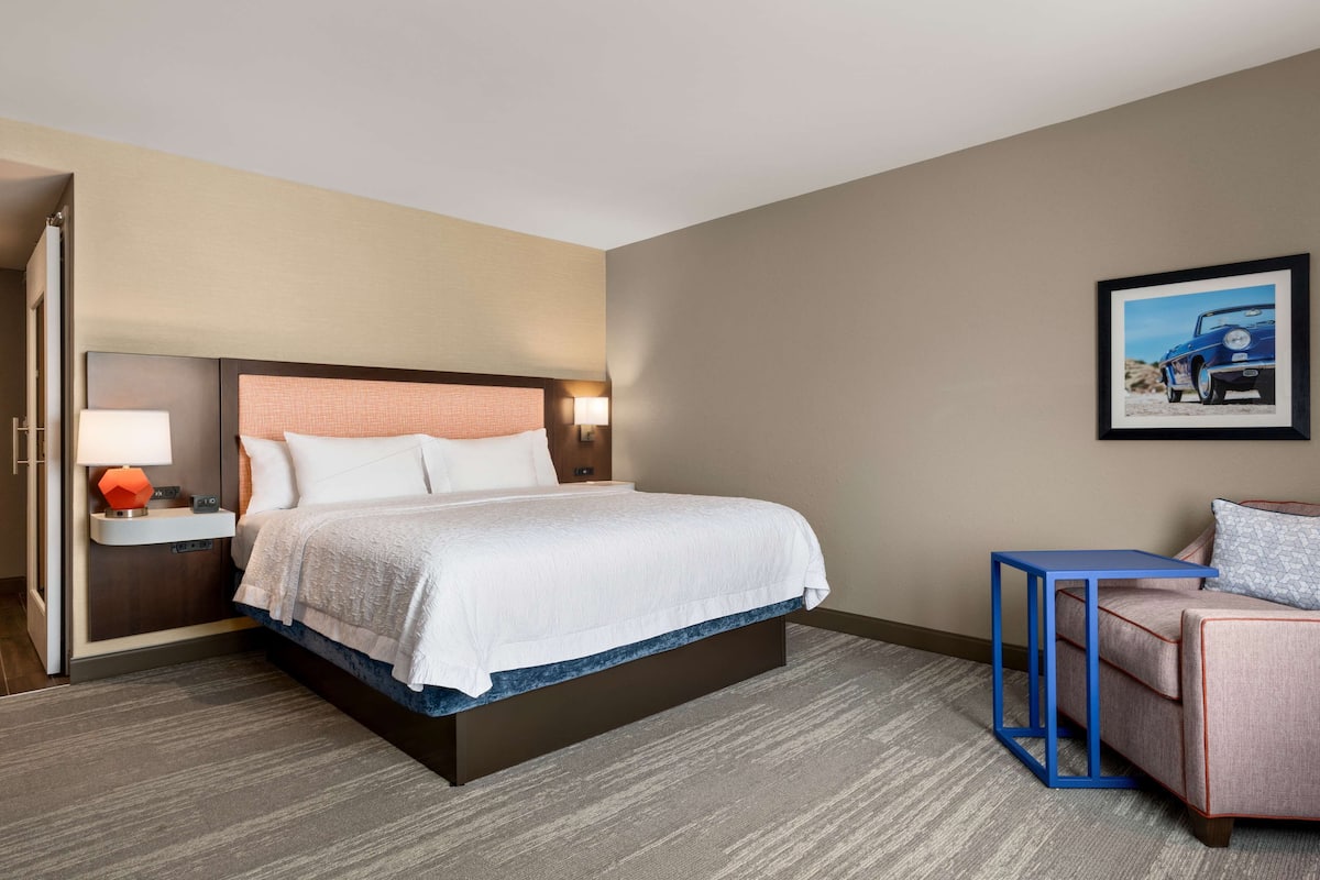 A hotel room with a large bed, a nightstand with a lamp, a chair, and a framed picture on the wall.