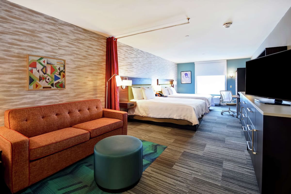 A hotel room with a sofa, a round ottoman, a bed, a desk, and a television.