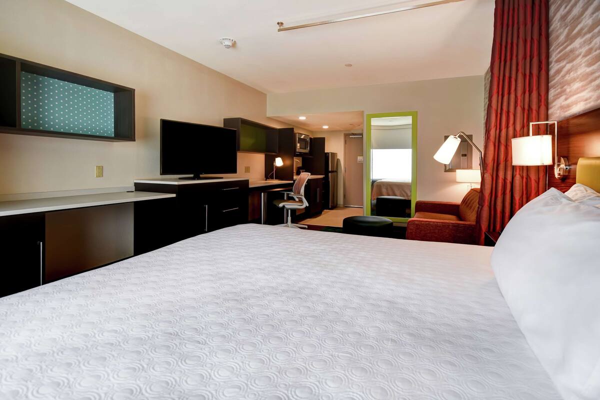 A hotel room with a large bed, a desk with a flat-screen TV, a chair, a small kitchenette, and a mirror.