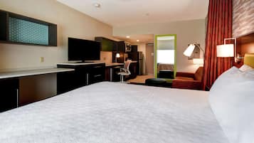 A hotel room with a large bed, a desk with a flat-screen TV, a chair, a small kitchenette, and a mirror.