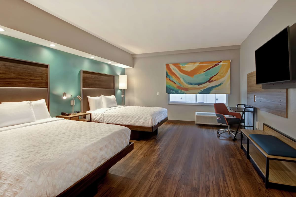 A hotel room with two beds, a desk, a chair, a TV, and a large abstract painting on the wall.