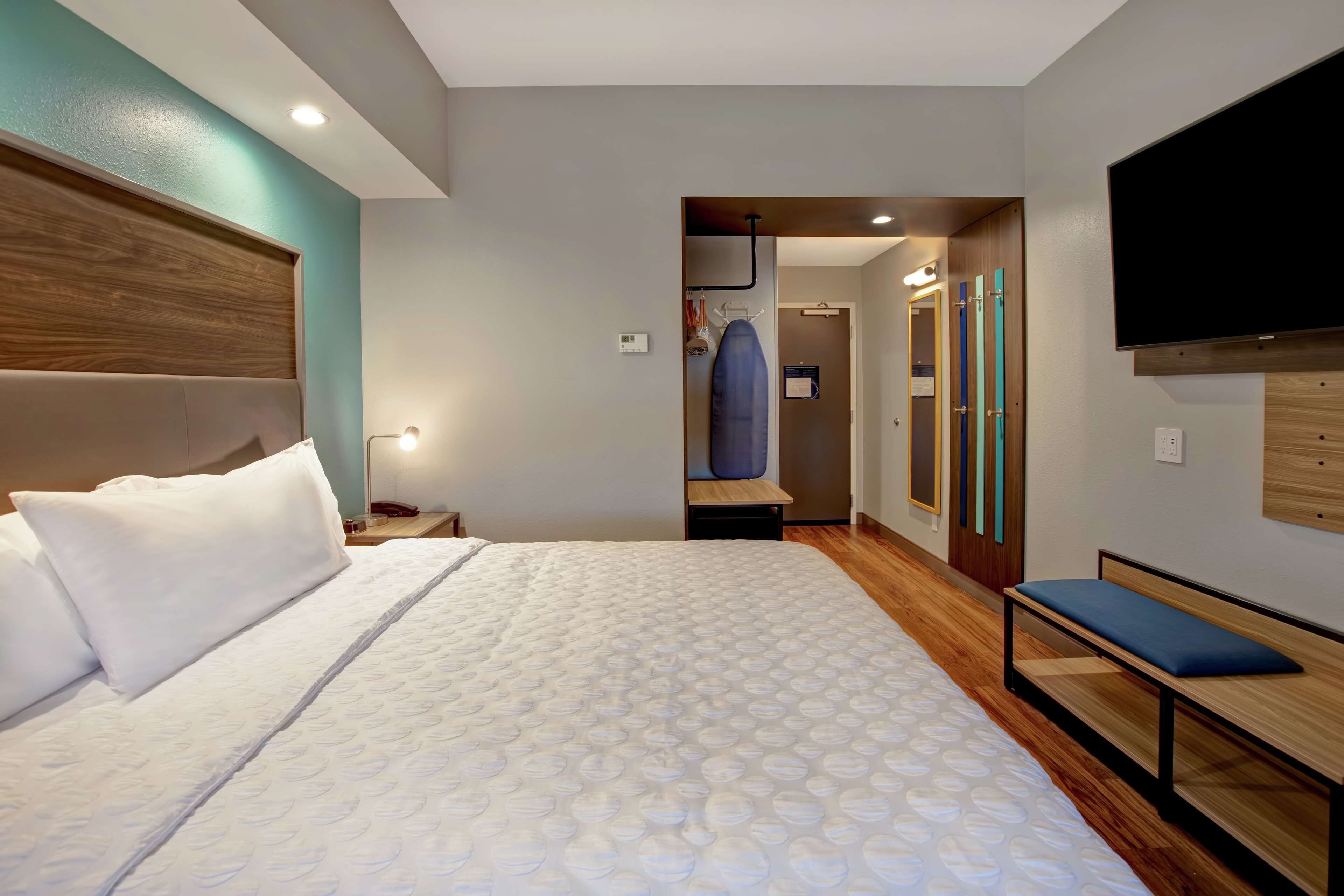 A modern hotel room with a large bed, a bench, a TV, and a walk-in closet.