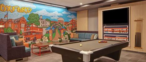 Una mesa de billar en una habitación con un mural de Oxford, un sofá y un televisor.