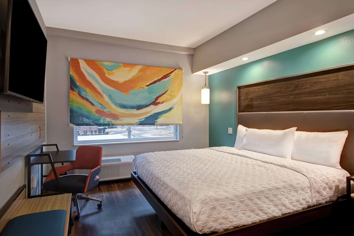 A hotel room with a large bed, a desk with a chair, a TV mounted on the wall, and a colorful abstract painting above the bed.