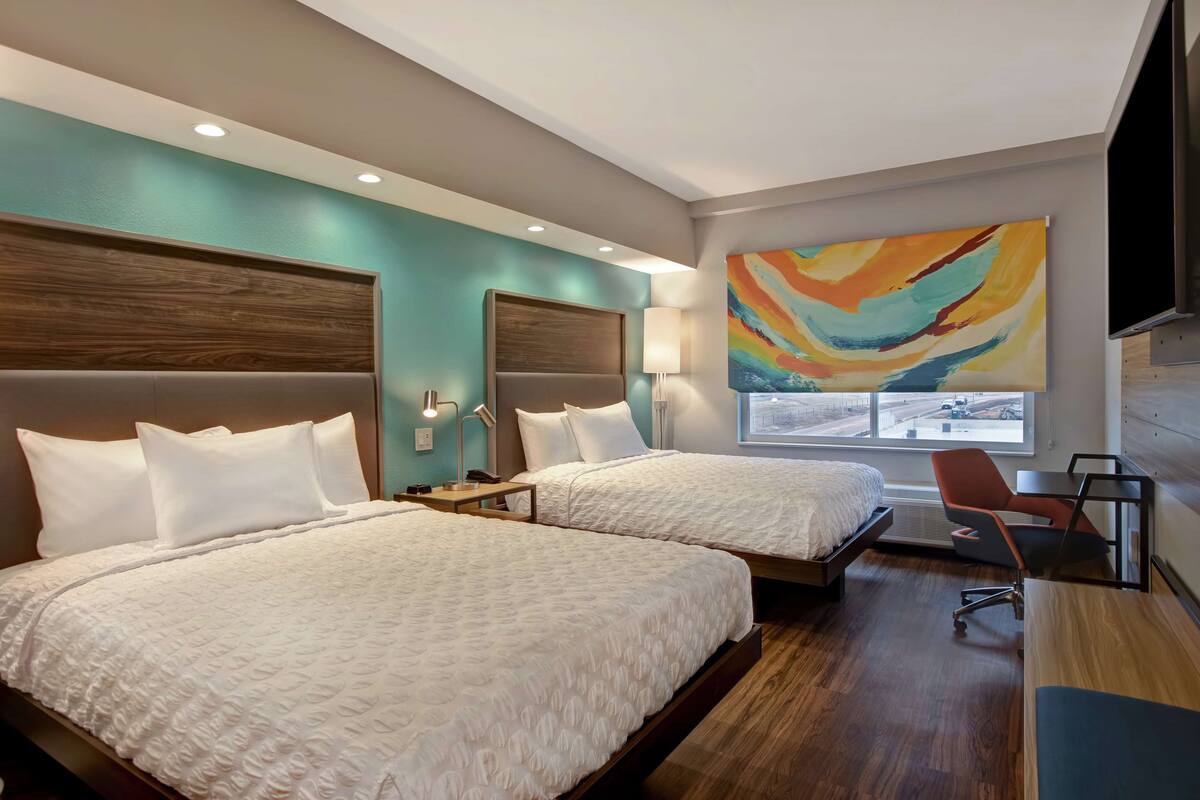 A hotel room with two beds, a desk, a chair, a TV, and a large window with a view.