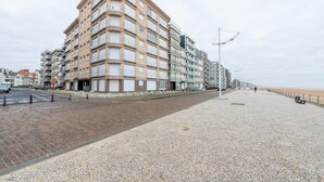 A coastal promenade with a row of multi-story buildings, a paved walkway, and a beach in the background. - Nevada 3-4 (Koksijde)