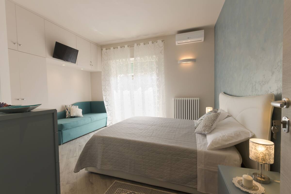 A bedroom with a bed, a TV mounted on the wall, a sofa, and a bedside table with a lamp.