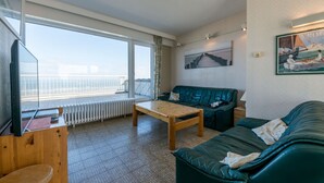 A living room with a large window, a wooden coffee table, a green sofa, and a television. - Magellan 701 (Koksijde)