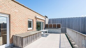 A rooftop patio with a wooden deck, a white table, and a view of a brick building. - Magellan 701 (Koksijde)