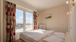 A room with two beds, a window with curtains, a radiator, a painting, and a wall-mounted light fixture. - Magellan 701 (Koksijde)