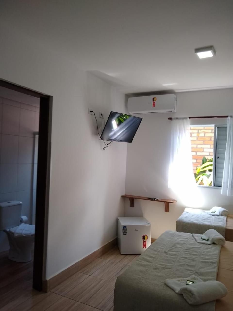 Quarto de hotel com cama, mesa de cabeceira pequena, televisão fixada na parede, ar condicionado e janela com cortinas.
