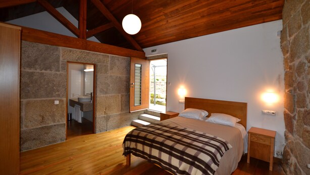 A bedroom with a wooden ceiling, stone walls, a bed with a plaid blanket, bedside lamps, and a view of the outside. - Casa de Santa Cristina (Marco de Canaveses)