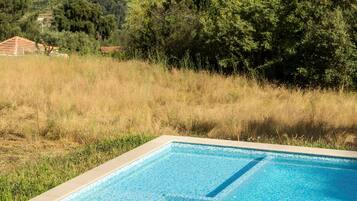 A rectangular swimming pool with clear blue water, surrounded by well-maintained grass and a few trees. In the background, there is a hilly landscape with scattered houses.