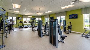 Een sportschool met diverse fitnessapparaten, grote ramen en een televisie aan de muur.