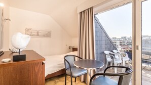 Ein Hotelzimmer mit einem Bett, einem Schreibtisch, zwei Stühlen, einem kleinen Tisch und Blick auf Gebäude durch ein großes Fenster.