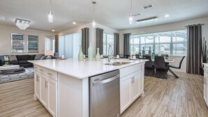 Een moderne keuken met een centraal eiland, witte kasten en roestvrijstalen apparatuur. Er is een zithoek met een tafel en stoelen, en een woonkamer met een bank en salontafel.