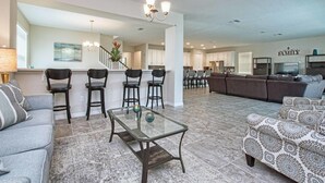 A spacious living room with a glass coffee table, a sofa, and a patterned ottoman. There is a kitchen area in the background with white cabinets and a dining area with chairs. - Solara 9 Bedroom House w Pool - 8100SA (Kissimmee)