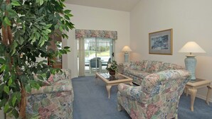 A living room with floral-patterned furniture, a glass-top coffee table, two lamps, and a large window with a view. - Greater Groves 4 Bedroom Home w Pool - 5204GG (Clermont)