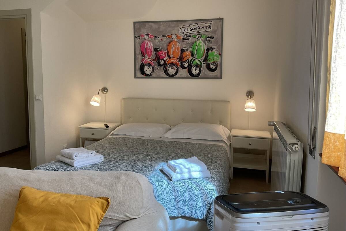 Une chambre à coucher comprenant un lit, deux tables de chevet, un radiateur, un tableau représentant des scooters accroché au mur, et une fenêtre avec des rideaux.
