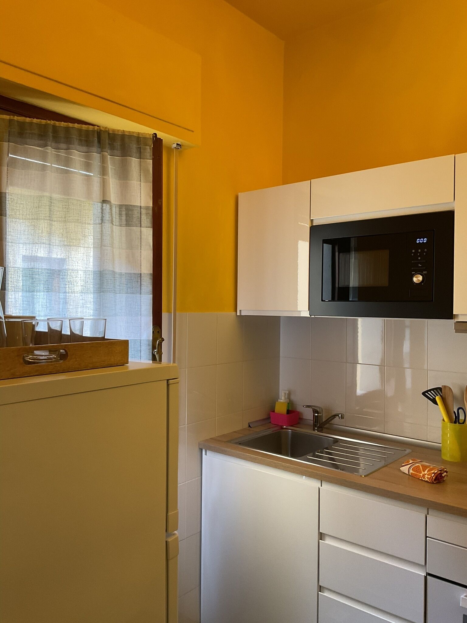 A kitchen with white cabinets, a microwave, a sink, and a window with a curtain.