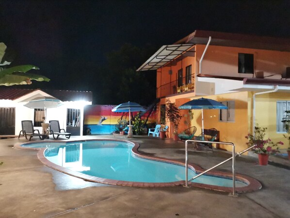Una piscina con sillas de descanso y sombrillas, rodeada de edificios con murales de colores y plantas en macetas.