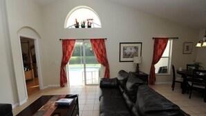 Ein Wohnzimmer mit schwarzem Ledersofa, Couchtisch und Essbereich mit Tisch und Stühlen. Ein großes Fenster mit roten Vorhängen, ein Bild an der Wand und eine Tür zu einem anderen Raum.