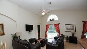 Ein Wohnzimmer mit zwei schwarzen Ledersofas, einem Flachbildfernseher, einem Deckenventilator und einem großen Fenster mit roten Vorhängen.