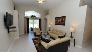 A spacious living room with a ceiling fan, a flat-screen TV mounted on the wall, a beige sofa, a black armchair, and a glass coffee table. - Indian Point 00 (Kissimmee)