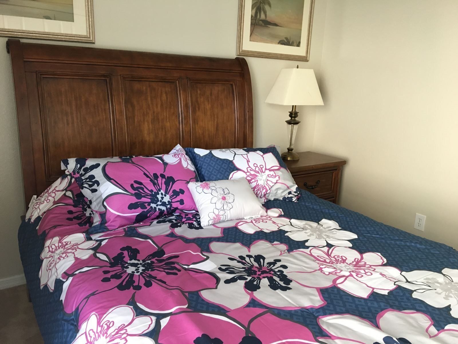 Una cama con ropa de cama floral, un cabecero de madera, una mesita de noche con una lámpara y una pared con un cuadro enmarcado.