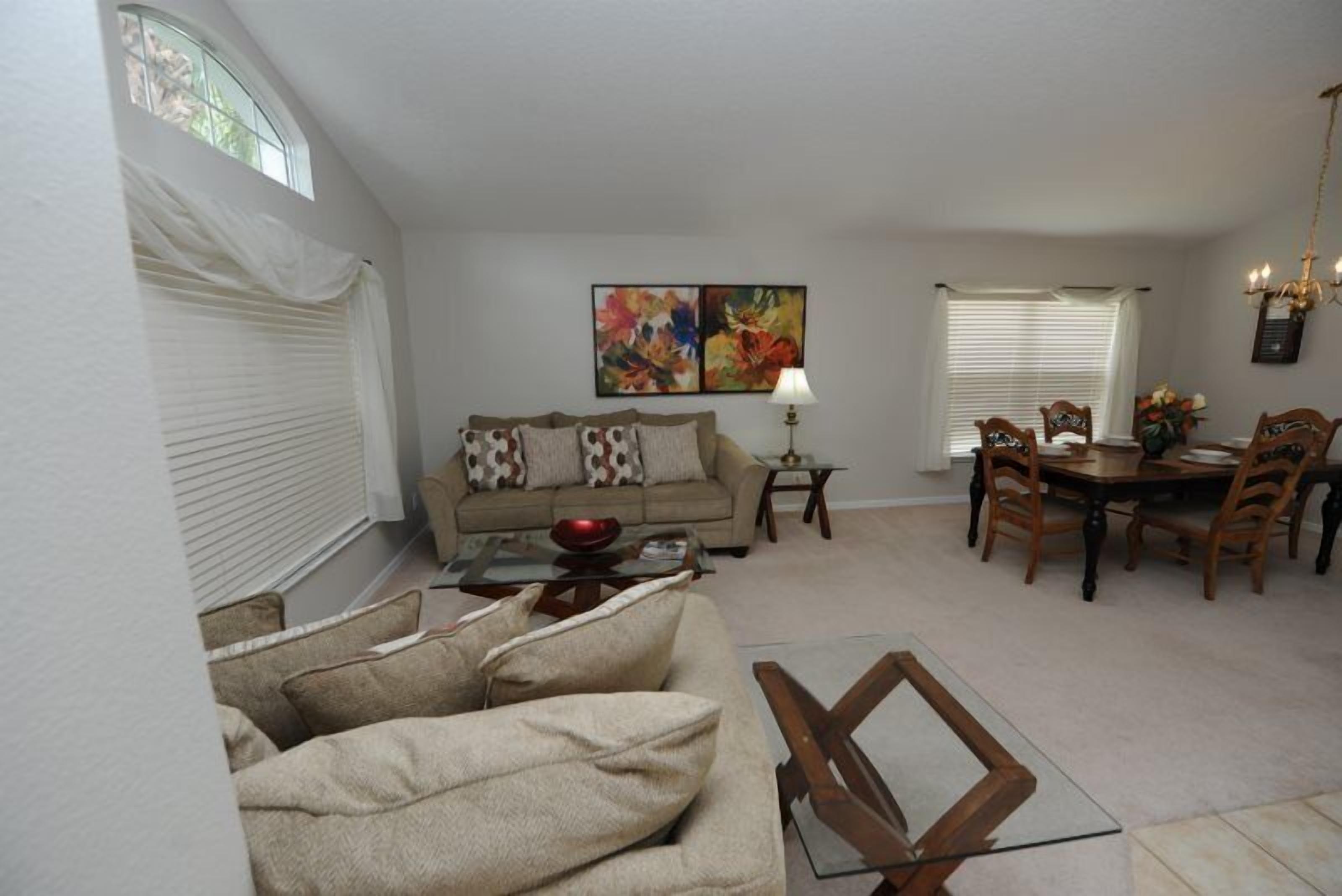 A living room with a sofa, coffee table, dining table, and chairs. There is a painting on the wall and a lamp on the table.