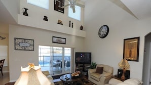 A spacious living room with a high ceiling, a television, a sofa, a coffee table, and a dining area with a view. - Cumbrian Lakes 25 (Kissimmee)