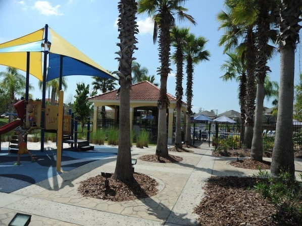A playground with a slide and climbing structures, surrounded by palm trees and a gazebo.