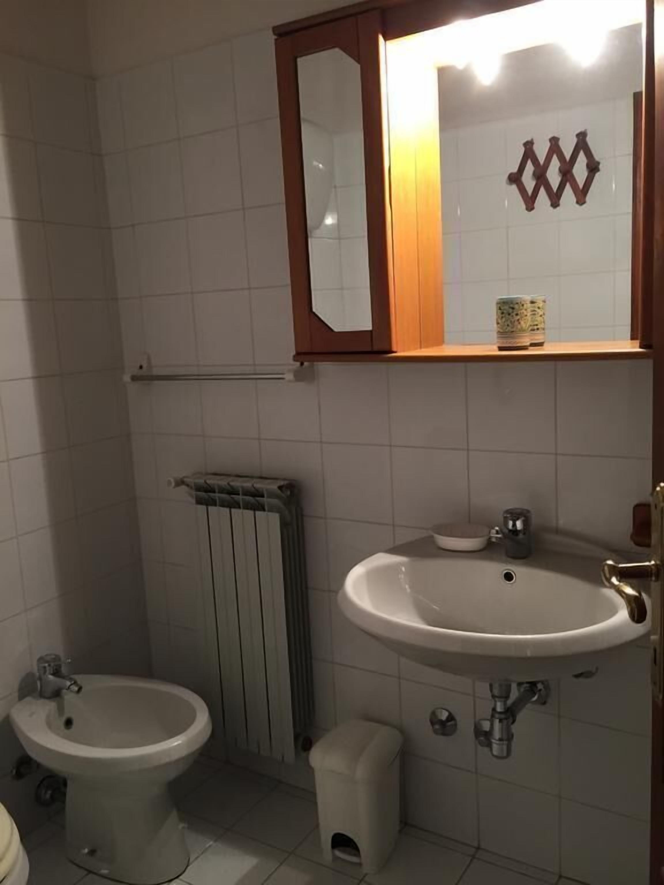 Un baño con lavamanos pedestal, dos inodoros, un radiador y un espejo sobre el lavamanos.