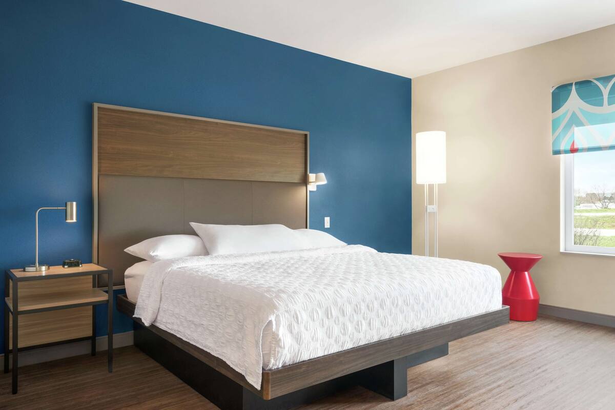 A modern hotel room with a wooden bed frame, a nightstand with a lamp, a red accent chair, and a window with a view.